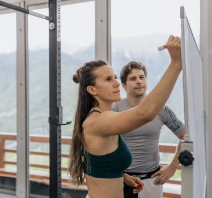 Female fitness coach writes on whiteboard as a male athlete watches in a gym.